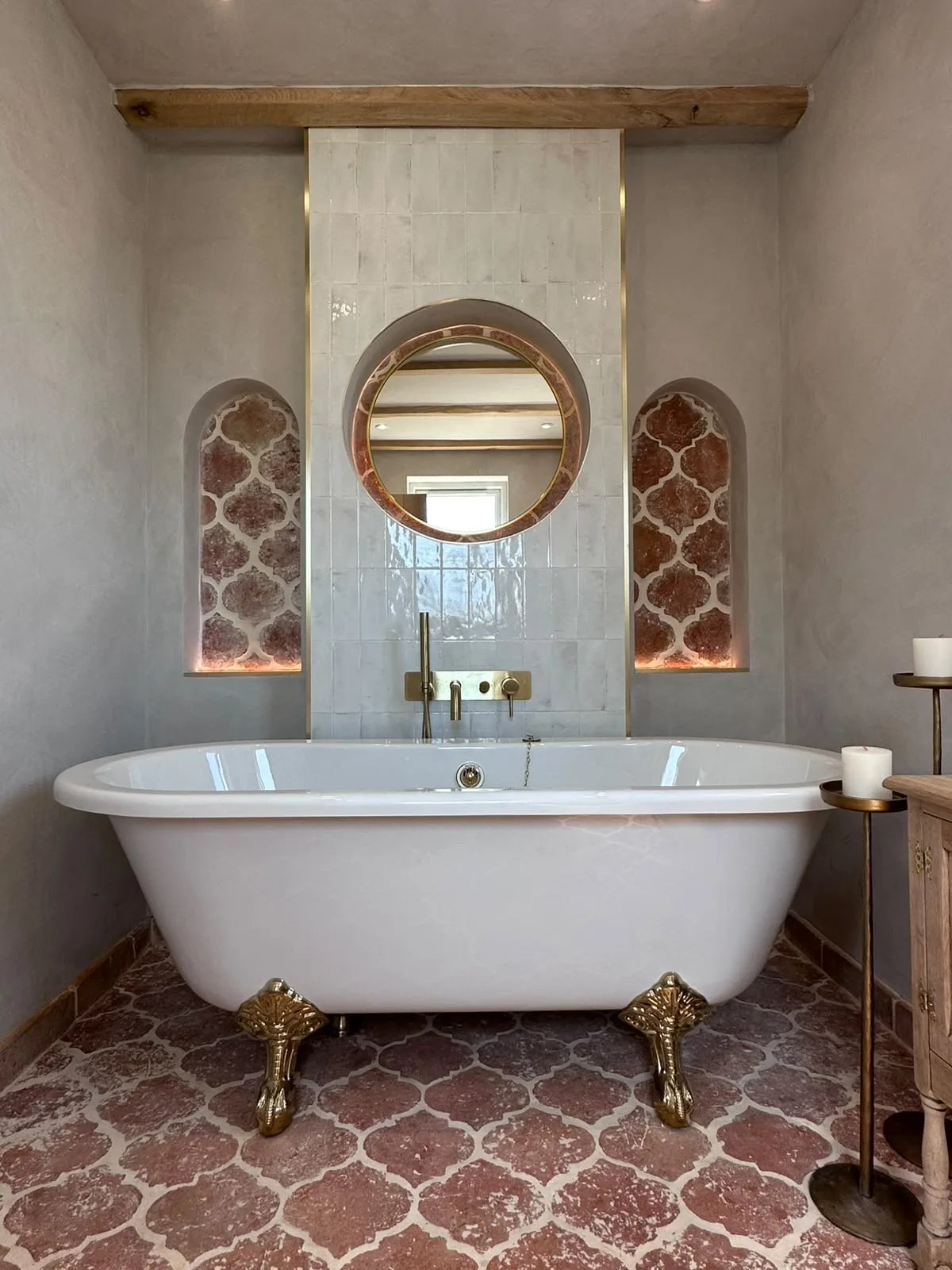 Rolltop bath with brass claw feet, terracotta floor tiles, arched tiled niches and a round brass mirror